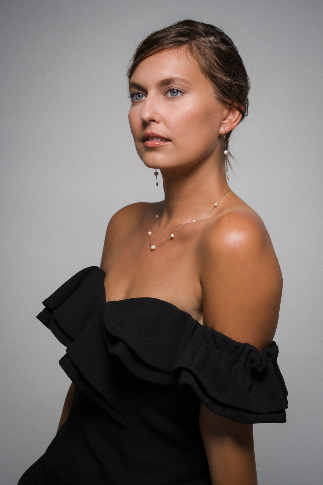 Woman wearing matching set of pearl jewellery, drop earrings and matching necklace, a black off-shoulder top against a gray background