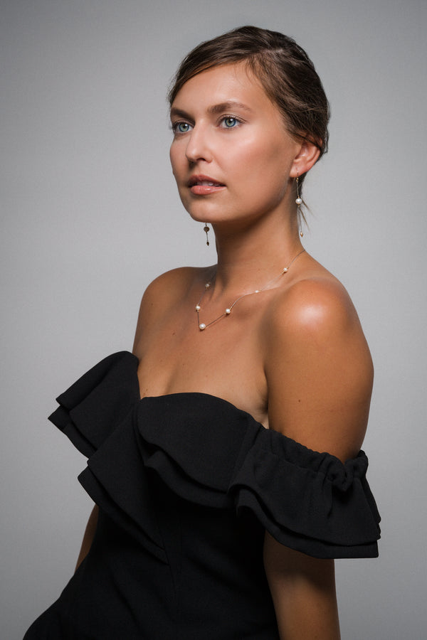 Woman wearing matching set of pearl jewellery, drop earrings and matching necklace, a black off-shoulder top against a gray background